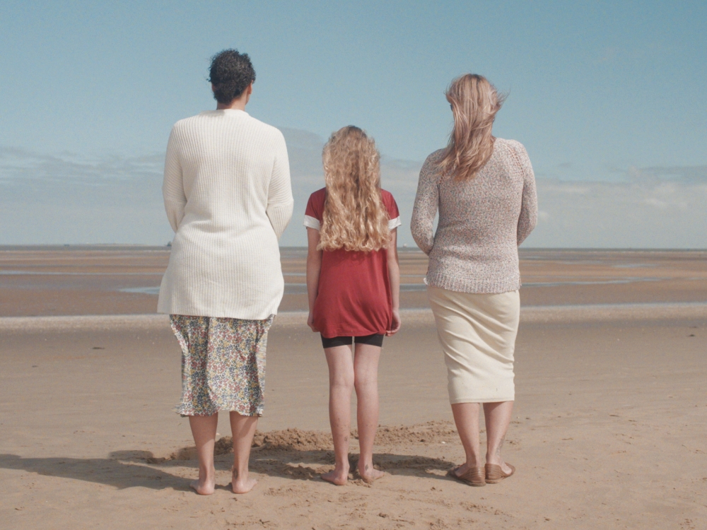 Any of us video final shot - three people standing on a beach their backs facing the camera. Left to Right - a lady with dark hair, a blonde haired girl and a blonde haired lady.